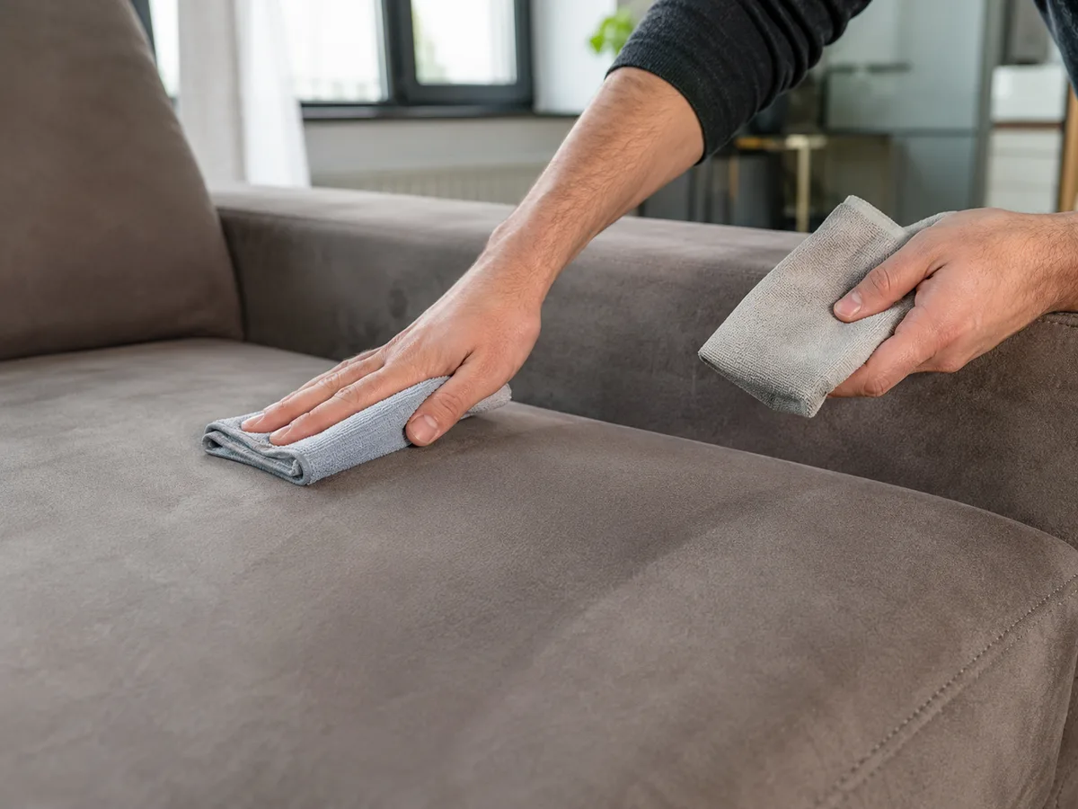 Mann arbeitet mit zwei Mikrofasertüchern parallel über die Sitzfläche eines braunen Alcantara-Sofas — gleichmäßig in eine Richtung