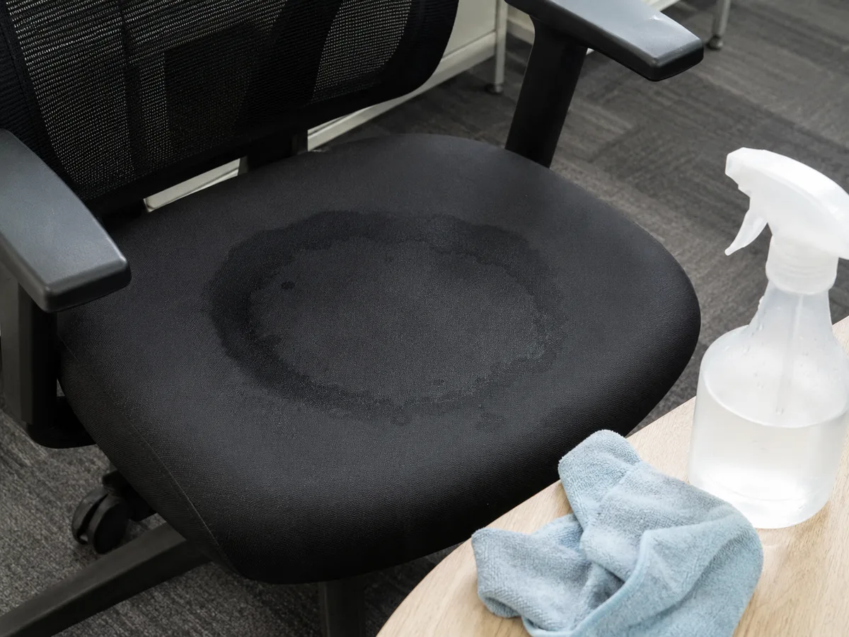 Schwarzer Bürostuhl mit feuchtem Wasserring auf der Sitzfläche, daneben Sprühflasche und blaues Tuch — Trocknungsphase