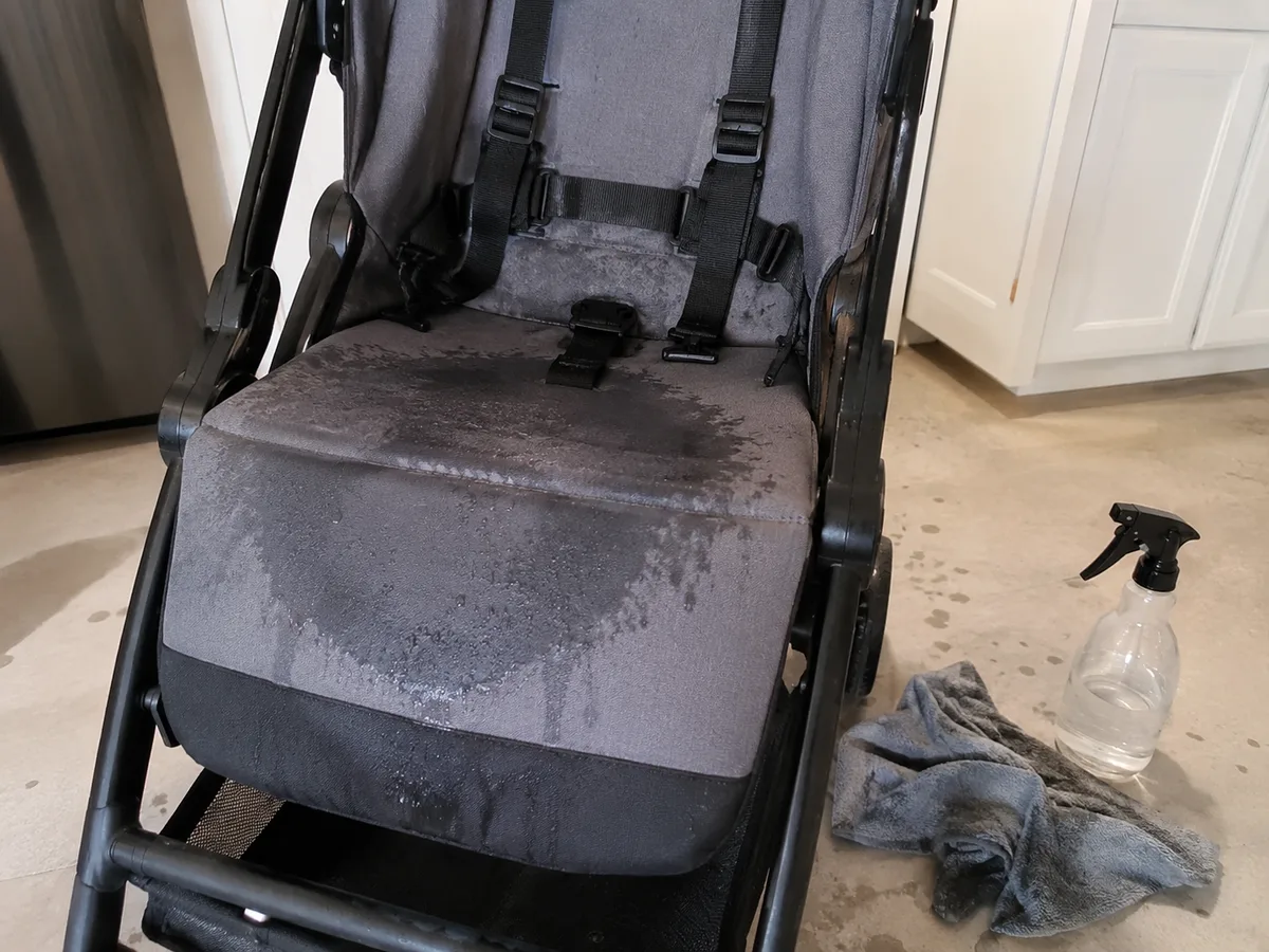 Sitzfläche eines Kinderwagens feucht nach der Reinigung, daneben Sprühflasche und grauer Lappen auf dem Boden