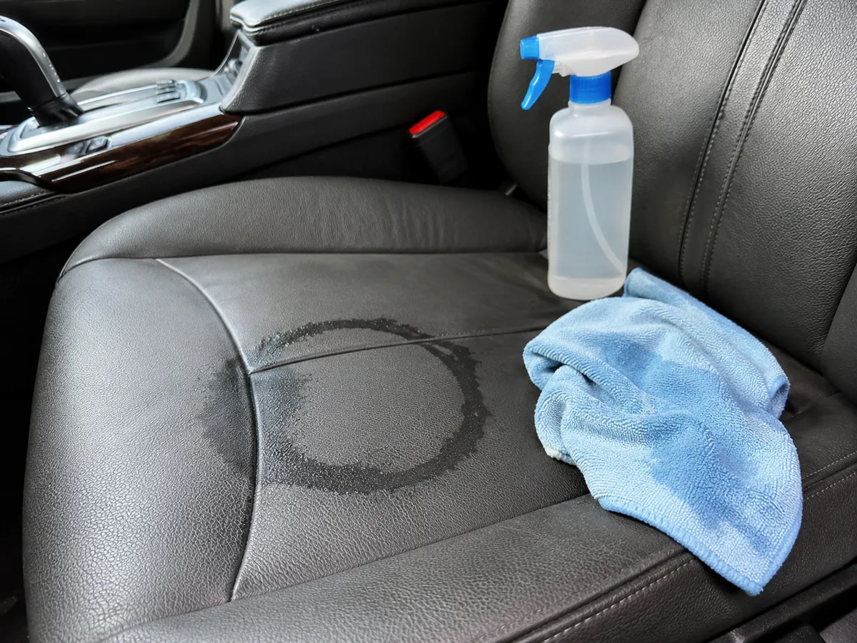 Dunkler Auto-Ledersitz mit deutlich sichtbarem Wasserring von einer Flecken-Behandlung, Sprühflasche und blaues Tuch daneben