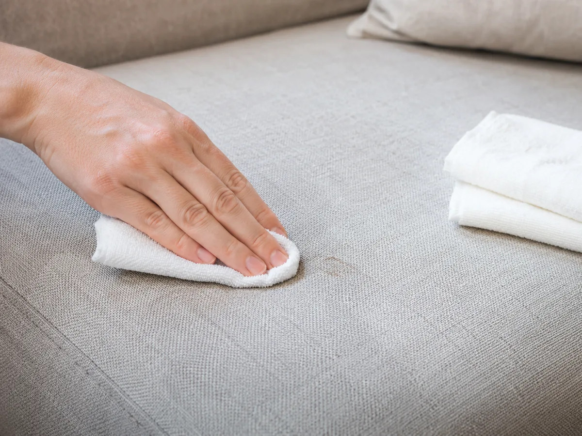 Hand tupft mit zusammengefaltetem weißem Tuch sanft auf einen kleinen feuchten Fleck auf einem hellen Stoffsofa, daneben weitere gefaltete Tücher