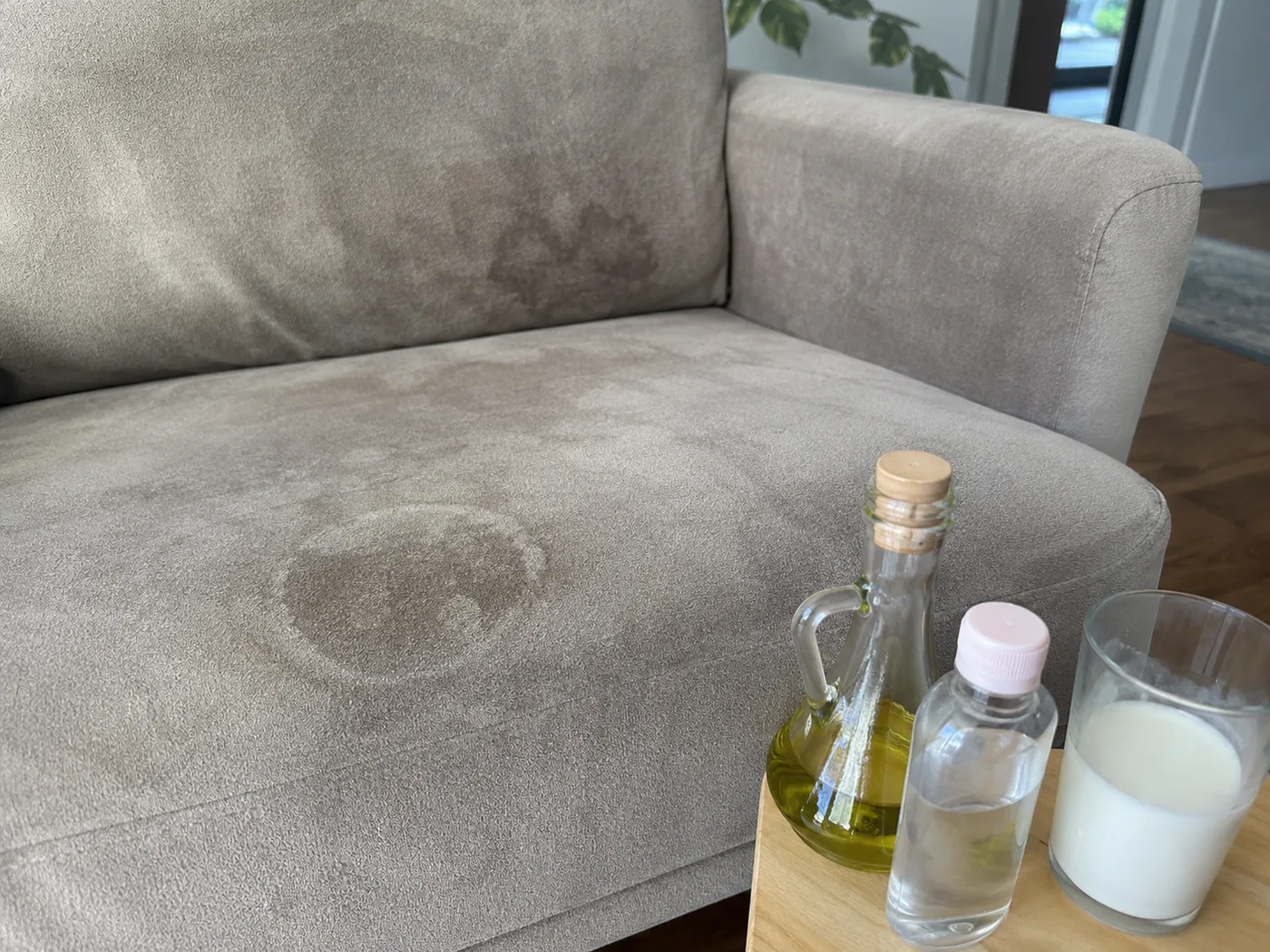 Drei Hausmittel-Flaschen — Olivenöl, klares Wasser und Milch — stehen auf einer Tablett-Ecke vor einem hellgrauen Velours-Sofa mit deutlich sichtbarem Wasserring auf der Sitzfläche